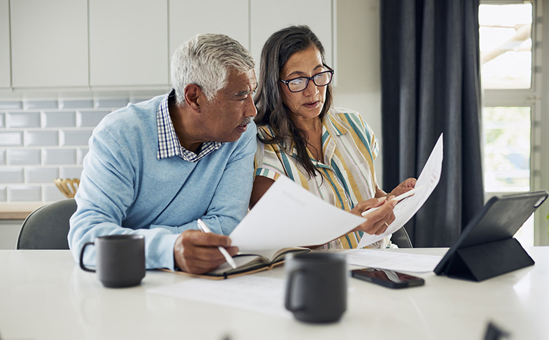 An older couple sits at their kitchen table reviewing financial documents. Year-end tax planning.